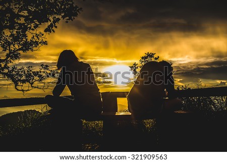 The silhouette of two women sitting next to each other on a single chair on the sunset background.