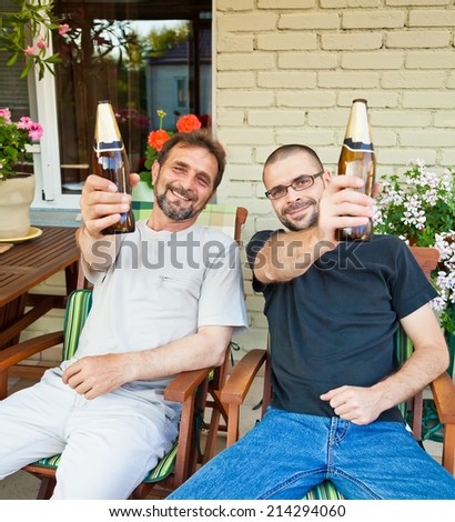 Two men - father and son - sitting in terrace with beer bottle cheering and smiling