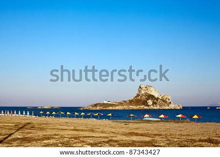 lined up beach chairs with parasols on empty beach of the Greek island Kos with view on the remote monastery on a little island Kastri
