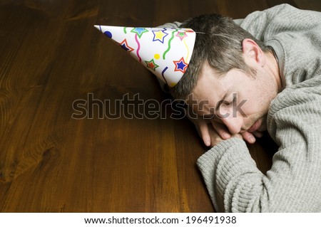 A man with a party hat is sleeping over a wooden table.