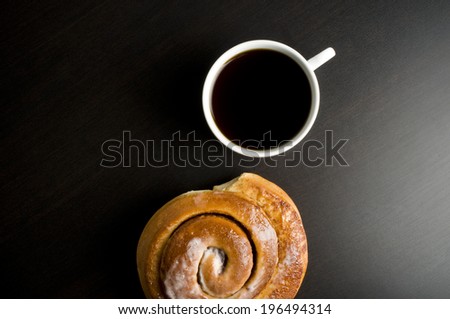 High angle perspective of a dark drink in a cup alongside a Chelsea bun.