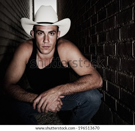 A man in a black tank top wearing a white cowboy hat crouching in an alley.