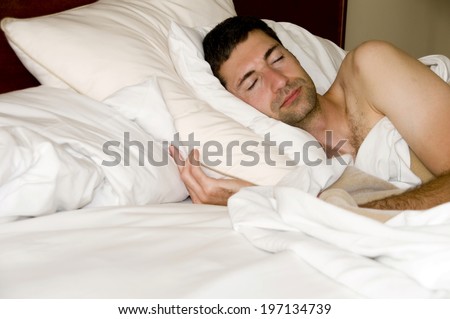 A man is sleeping topless on a few white pillows and under white sheets.