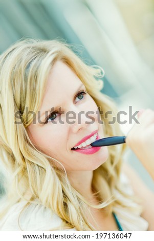 A smiling blonde woman with hazel eyes biting a pen and looking away.