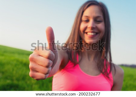 Happy woman giving thumbs up success hand sign outside on field smiling joyful. Sporty fit young Caucasian female fitness model outdoors.
