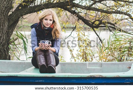 Young blonde woman resting in an old boat and holding a cup of coffee or tea.
