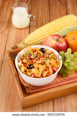Corn flakes bowl in tray with apple, orange, grapes and corn with milk jar on wooden background