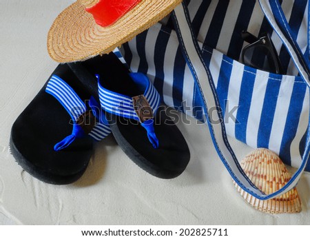 Summer scene of beach hat, beach bag and flip flops with blue and white stripes. The straw hat has a red band. The items are on clean, white sand. A seashell and sunglasses complete the scene.