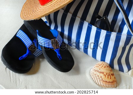 Summer scene of beach hat, beach bag and flip flops.  The beach bag and the flip flops have blue and white stripes.  There is a straw hat with a red band and a seashell in the sand beside the bag.