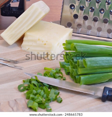 Ingredients and tools of cheese fondue include swiss and fontina cheeses, a cheese grater, chopped green onions and fondue forks all on a cutting board with a fondue pot in the background