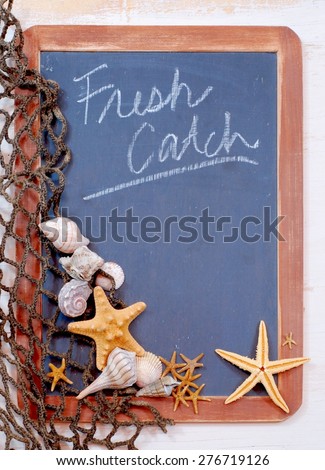 Seafood menu image of blackboard with fishnet and seashells and starfish gathered on left. The slate on the blackboard is rough and the brown frame is worn. Message is fresh catch with copy space