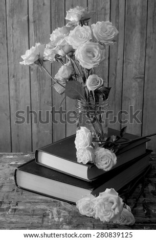 Black and white image of tired, white miniature tea roses in a mason jar on a stack of dusty books. The table top is old wood with many paint stains. Background is rustic wood. Side lighting from left