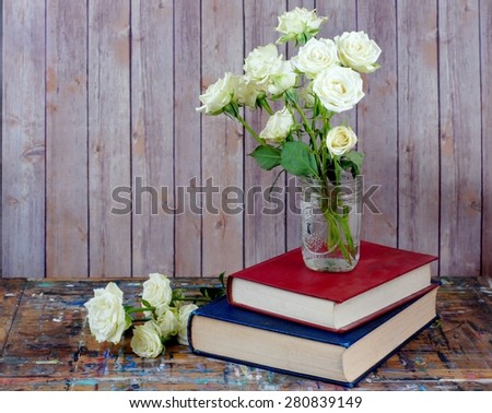 Tired, white miniature tea roses in a mason jar on a stack of dusty books. The table top is old wood with many paint stains. Background is rustic wood with copy space. The books are red and blue.