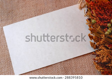 Blank message card of a parchment paper card tucked into a fall arrangement of dried leaves and grasses making border on the right with burlap background. Image has seasonally appropriate warm tones.