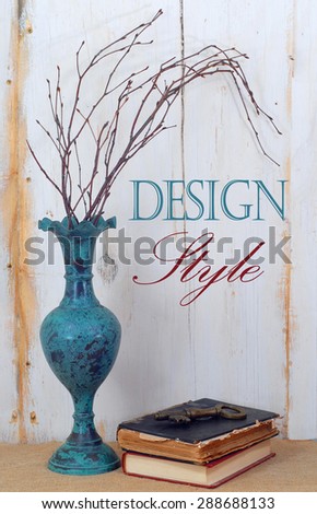 Greened copper looking vase with dried twiggy branches in front of a rustic wooden wall with worn white paint. Old books and an antique key lay on burlap in a grunge or shabby chic style. Text added.