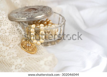 An old crystal jewelry box with the aged silver lid off contains strands or pearls and a gold and diamond locket. All items are on soft cotton and delicate lace fabric in white and ivory. Copy space.