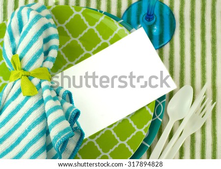 A casual place setting in green and turquoise with fun shapes on plates and white plastic utensils. Striped towel background. Cloth napkin tied with green ribbon. Card blank for text. Overhead view.