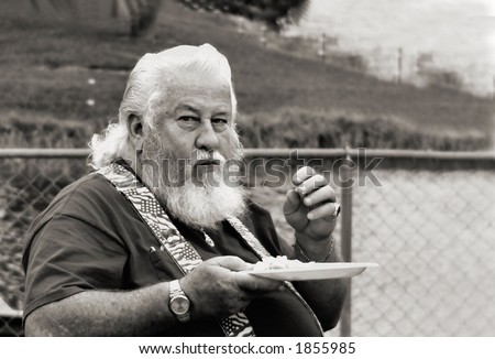 Black and white elderly overweight man sitting by a chain link fence next to a grassy bank by a lake eating with unhappy expression. The man, who is wearing suspenders, has long white hair and beard