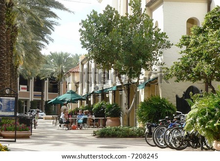Charming city scene in trendy restaurant area. Viewer is looking down a walkway in front of shops with landscaping, umbrellas and awnings. There are bicycles parked on the right. People sit at a table