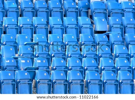 Empty bright blue stadium seats create repeating pattern. Horizontal image is filled with folded seats with one seat being unfolded, in the upper right. Six rows of eight chairs each.