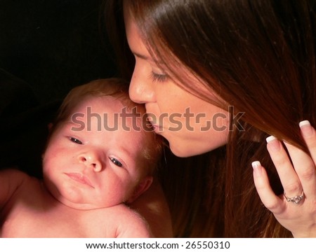 A beautiful young mother kissing her infant on the head. She has shiny brown hair. The baby\'s eyes are open. The mother is holding her hair back with one manicured hand. Isolated on black.