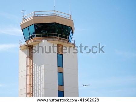 Aircraft control tower on sunny day. The tower is large with windows all around.  There are a few high clouds and a distant airplane is taking off lower right. Horizontal composition leave copy space
