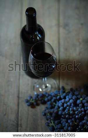 An opened bottle and glass of red wine with ripe, dark blue bunches of wine grapes on old, wooden background.