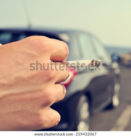 closeup of a man with a burning cigarette in his hand while is waiting besides a car parked next to a no traffic road