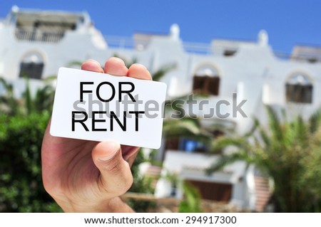 closeup of the hand of a man holding a signboard with the text for rent written in it in front of a house