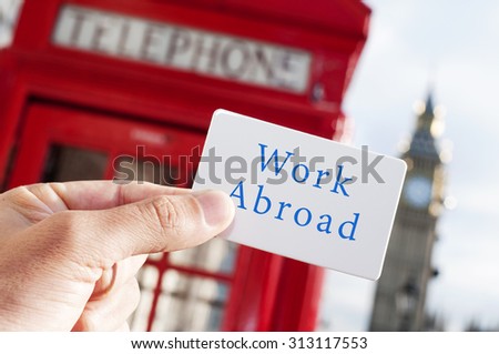 the hand of a young man showing a signboard with the text work abroad, with a red telephone booth and the Big Ben in the background, in London, UK