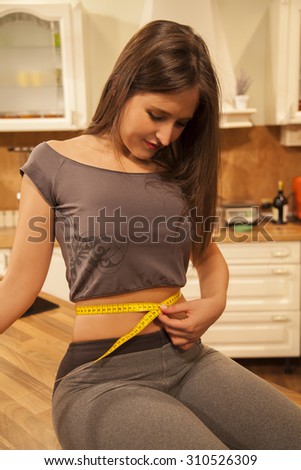 Smiling young woman dieting and measuring her waist with measuring tape at home.