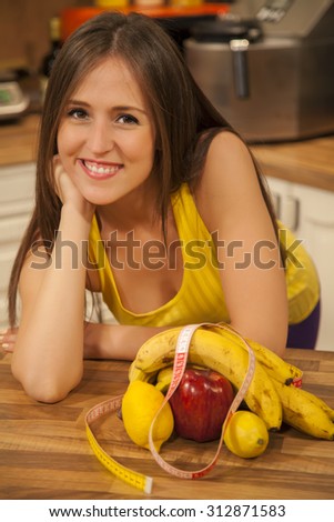 Smiling young woman dieting. She is leaning on the kitchen counter in front of fruits with tape measure and looking at camera.