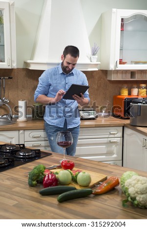 Young man leaning on the kitchen counter at home and using touchpad. He is taking a break from cooking. On the kitchen counter there are cutting board with glass of red wine, fresh vegetables