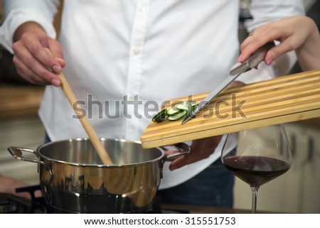 Close-up of a woman assisting a man in food preparation and adding sliced zucchini to cooking pot.
