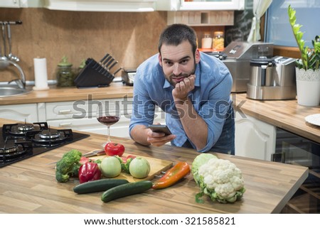 Young man using smart phone in the kitchen. He is leaning on kitchen counter and text messaging while looking at camera.