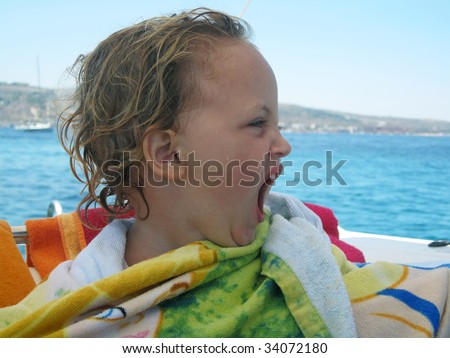 a young caucasian toddler sitting on a boat in the middle of the sea wrapped in a towel and  yawning