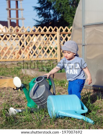 little boy plays with a watering can in the garden, next to the greenhouse