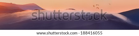 Majestic panorama of mountains at sunrise with pack of flying birds.