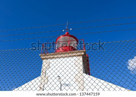 Bright Lighthouse beyond chain link fence against Blue Sky Background
