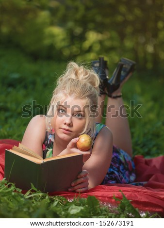 The girl lays on a glade, reads the book and eats an apple