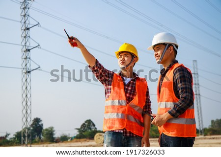 Asian engineer manager and foreman or leader discussion and pointing to construction site project on workplace and High voltage power line pylon in the background. Teamwork, Leadership concept.