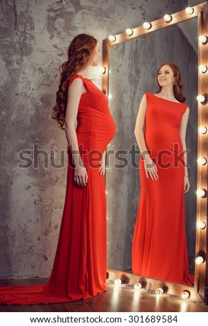 Pregnant girl with red curls standing in red dress and looks in a mirror