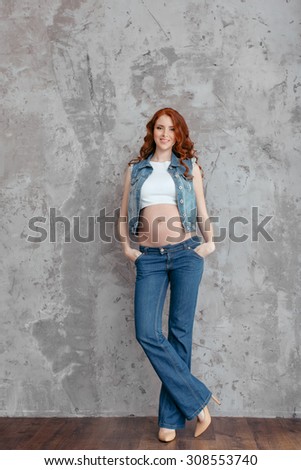 Studio portrait of beautiful slim sexy pregnant woman wearing casual jeans over grunge gray wall