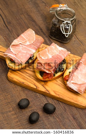 Snack with raw ham and black olive paste on white bread in vertical format
