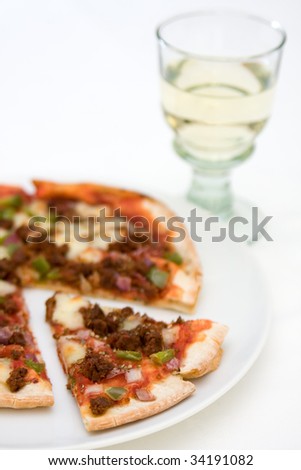 A pizza with spiced beef, green peppers and red onion on a white plate with a slice cut out and glass of white wine in the background, on a white tablecloth