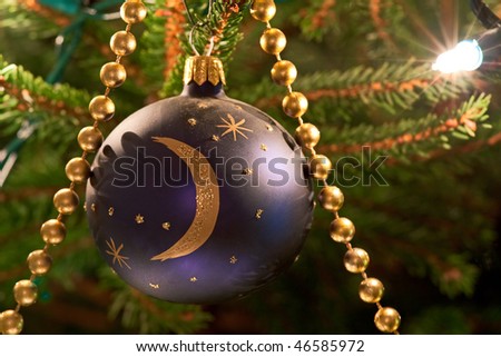 A blue and gold bauble hangs on a tree as a Christmas decoration, with gold beads and single fairy light. Photo has short depth of field.