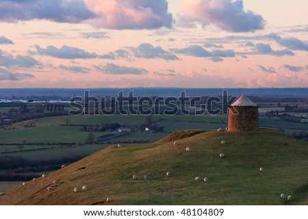 View of a country park with a stone monument, also a \'trig point\', and sheep grazing on the hillside, and cloudy pink sunset sky in the background.