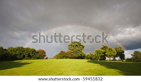 A rainbow with a hint of a double rainbow appears over a lush apple tree orchard in a domestic garden in late summer.