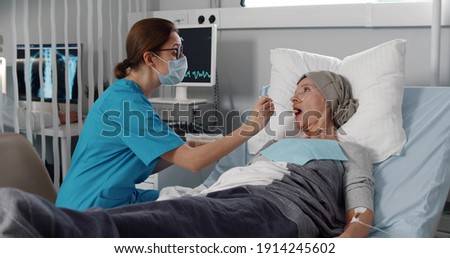 Nurse feeding mature woman having cancer with porridge. Medical worker in safety mask and scrubs feeding ill senior female patient lying in bed after chemotherapy