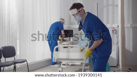 Janitors in scrubs and safety mask cleaning hospital room. Medical staff in protective wear wiping floor and changing bedsheets in empty ward in clinic